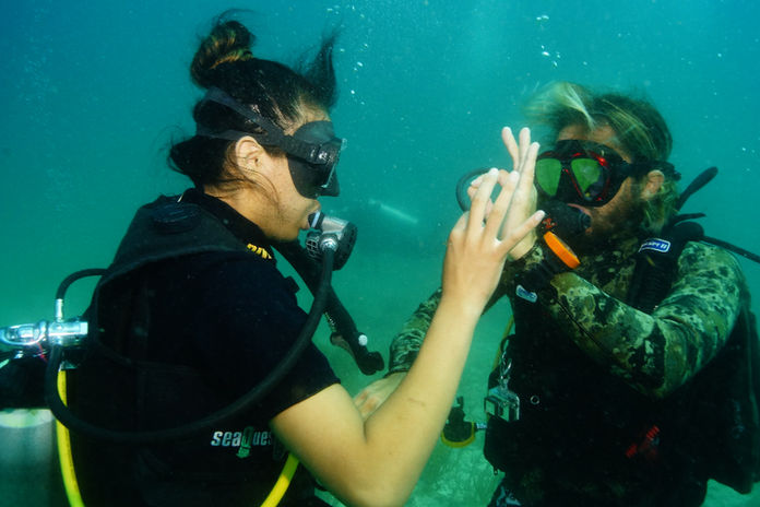 Two scuba divers underwater, practicing hand signals. Water is clear blue-green. One in camo gear, both in black masks, during Rescue Diver Cancun training.