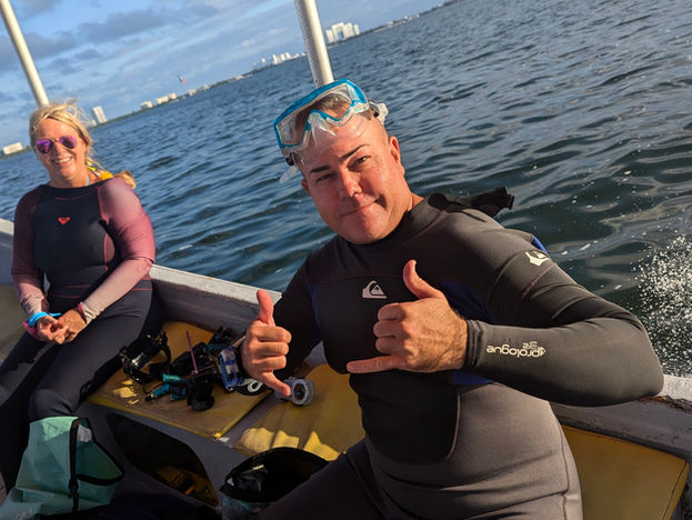 Two people in wetsuits on a boat; one gives a shaka sign. Sunny day on the water with a city skyline in the background.