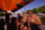 Smiling men on a dive boat under an orange canopy, one making a shaka sign, with marina and boats behind.