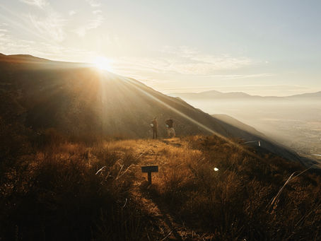Two hikers stand on a sunlit hillside at sunrise, surrounded by dry grass and mountains. A small sign is visible in the golden light.