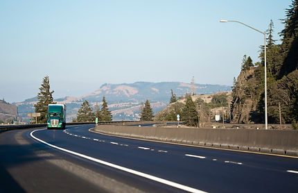 Green semi-truck driving on a curved highway with mountains and trees in the background. Clear blue sky and relaxed mood.