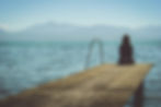Person sitting on a wooden dock overlooking a calm lake with mountains in the background. Clear blue sky, serene and contemplative mood.