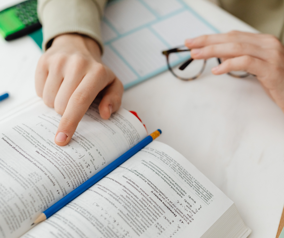 Hands point at text in an open book on a white table. A blue pencil lies across the pages. Glasses and a lined notepad are nearby.