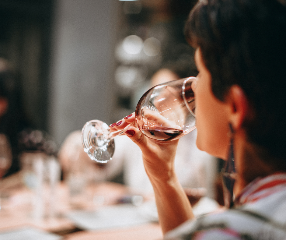 Person sipping red wine from a glass, blurred background with soft lighting, creating a relaxed, intimate setting.