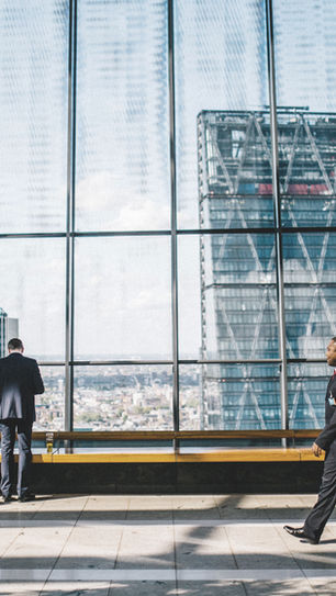Business people in a modern glass building, with cityscape views outside. A man walks while others converse or look outdoors. Bright daylight.