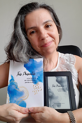 Uma foto minha: mulher branca, cabelos grisalhos, sorrindo, segurando duas versões do meu livro publicado intitulado "Três presentes: uma história de infertilidade, perda e vitória". À esquerda a cópia física, com capa branca com detalhes em aquarela azul e linhas douradas e à direita a mesma capa mas na versão para o kindle, em preto e branco.