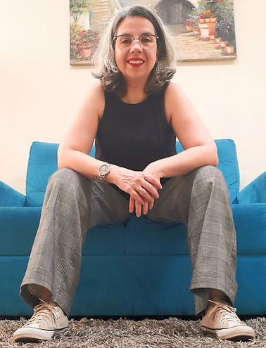 Uma foto minha: mulher branca, com cabelos grisalhos, de mãos unidas, sentada em um sofá.