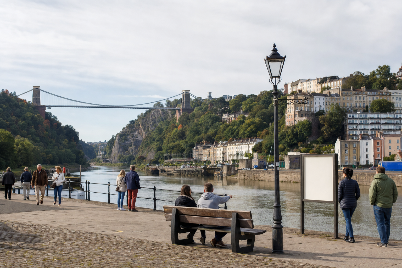 People walking along the waterfront with Clifton Suspension Bridge and hillside houses in the background.