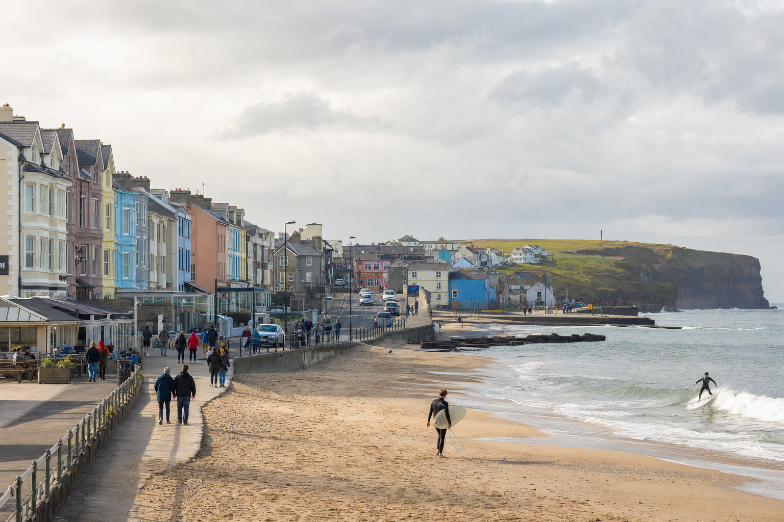 A coastal town with colourful buildings, people walking along the promenade, and a surfer riding the waves in the sea.