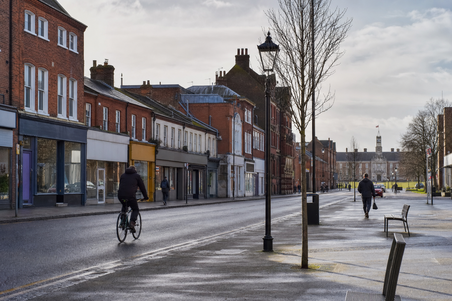 A cyclist and pedestrian on a quiet, overcast high street lined with brick buildings and leafless trees.
