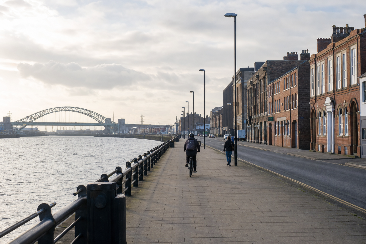 A person cycles along a riverside path beside historic buildings and a bridge in the distance under a cloudy sky.