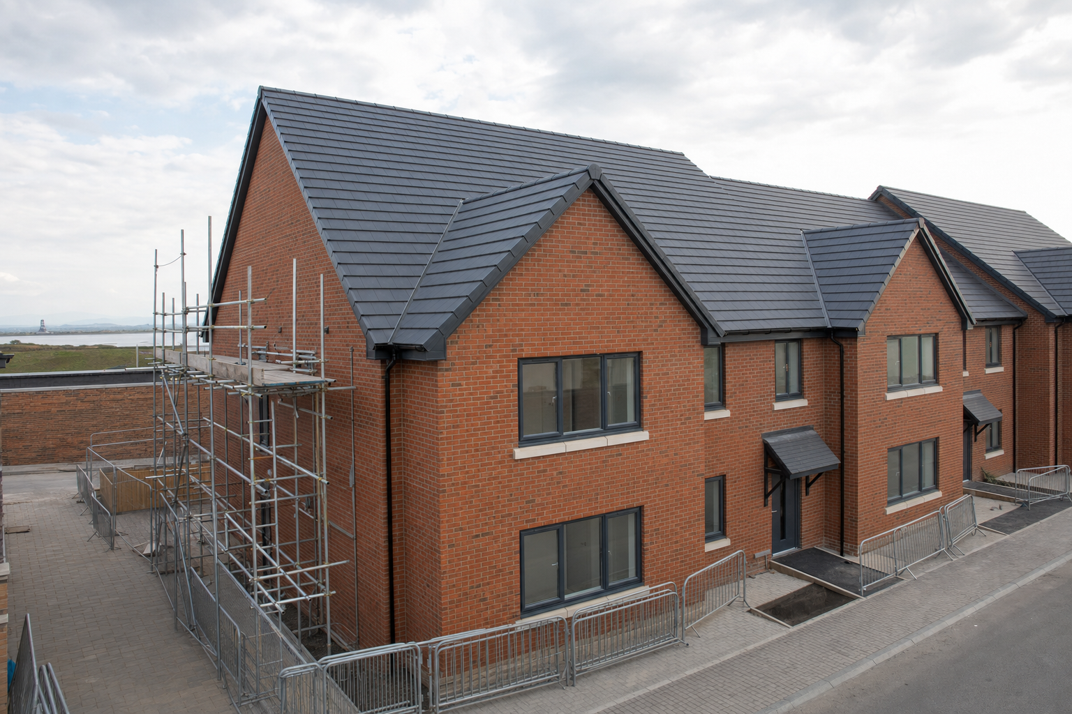 Newly constructed red brick houses with scaffolding and fenced pathways under a cloudy sky.