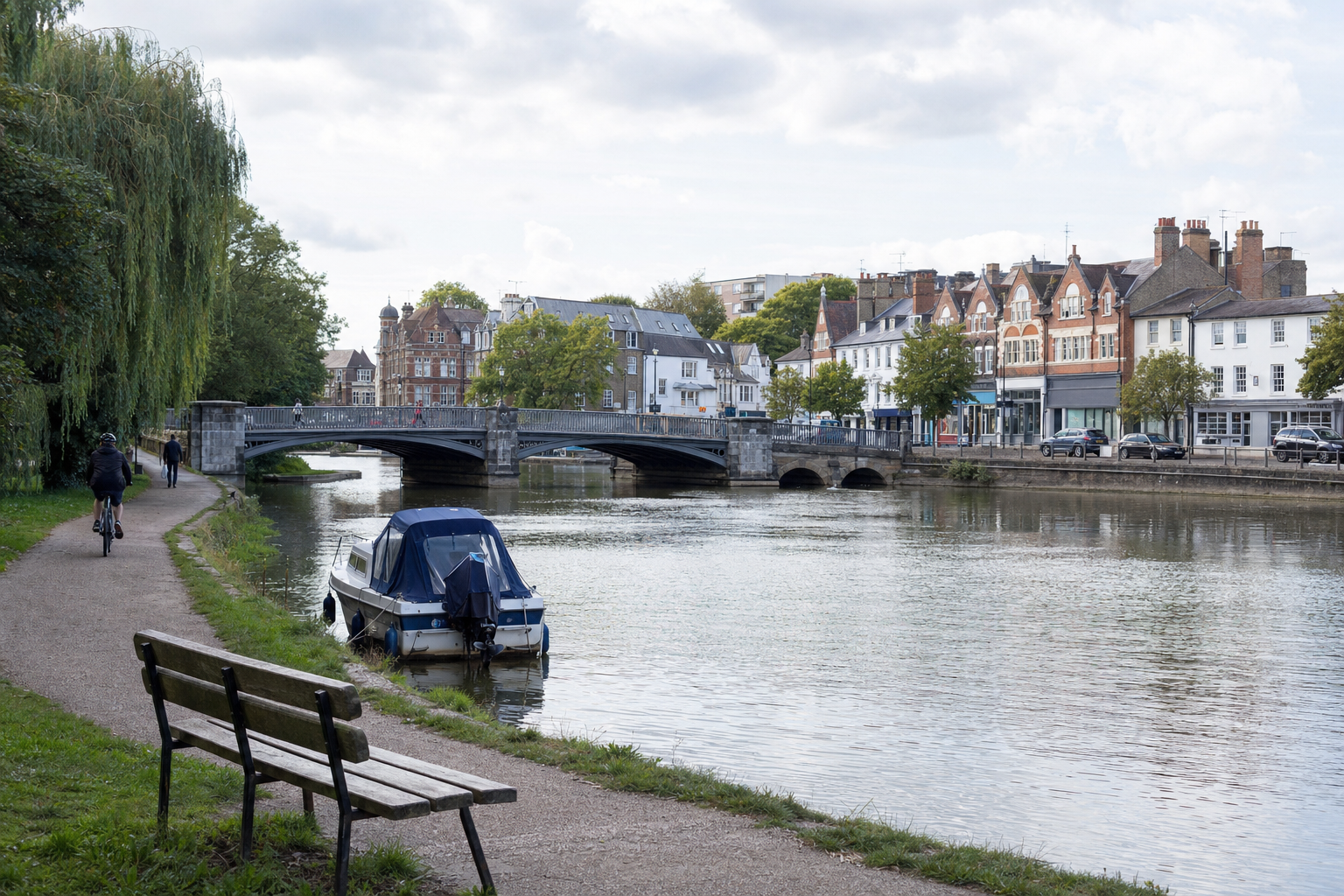 Canal-side scene with a moored boat, path, cyclists, and a stone bridge leading to charming townhouses.