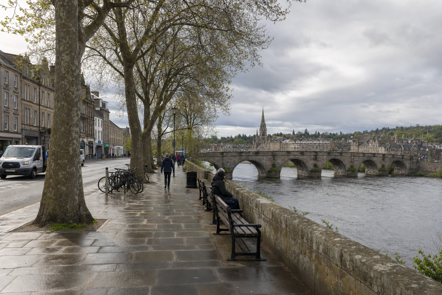 Riverside path with benches and pedestrians, offering a view of a historic stone bridge and buildings under a cloudy sky.