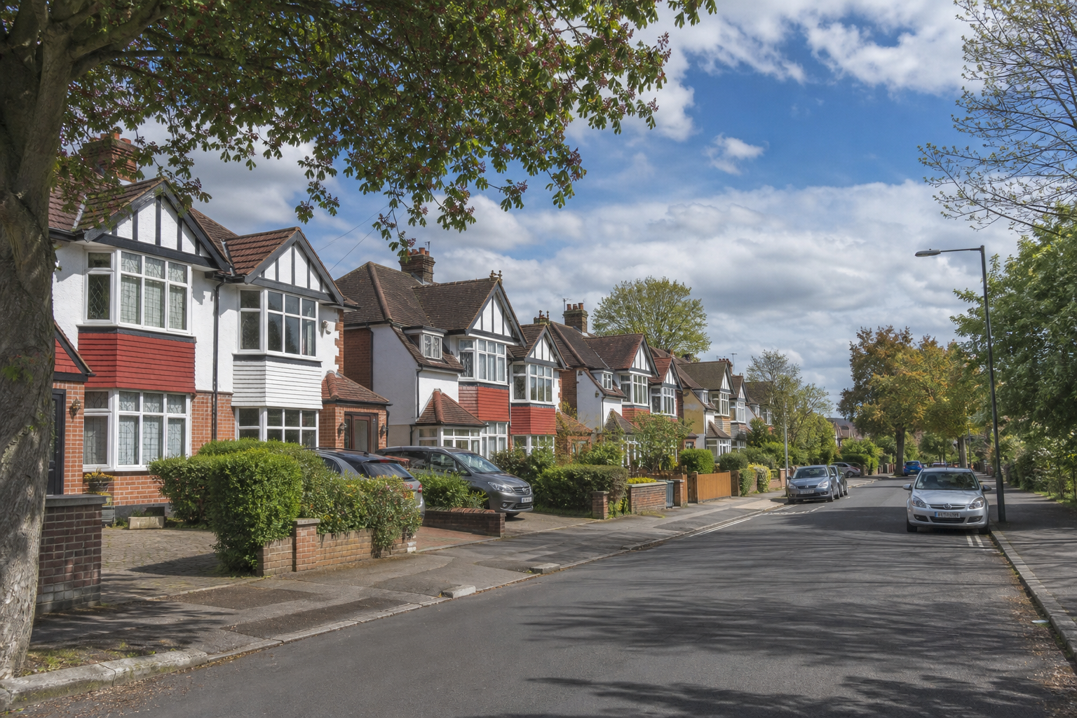 Suburban street lined with traditional houses, greenery, and parked cars under a partly cloudy sky.
