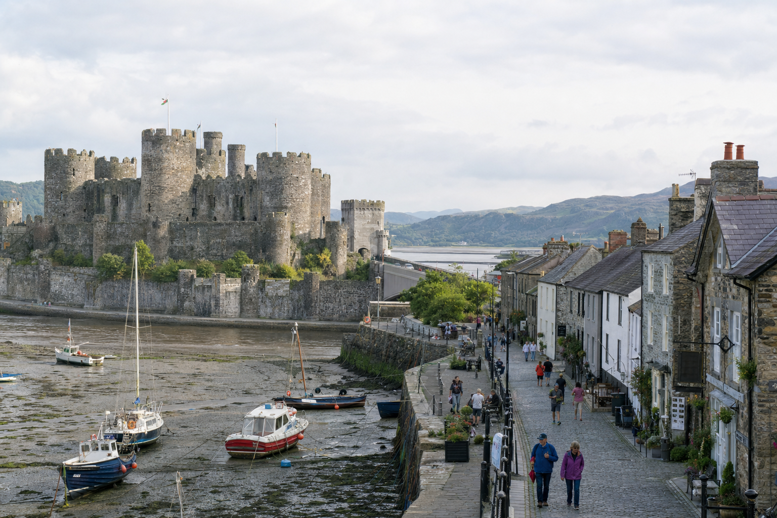 Medieval castle overlooking boats in a tidal river with a cobblestone street and people walking beside stone buildings.
