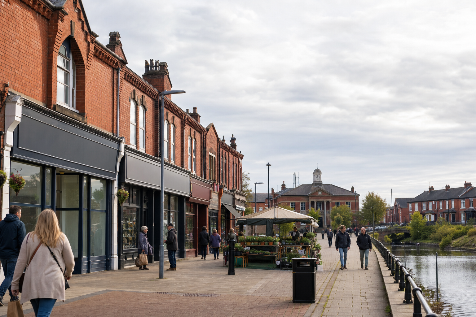 People walking along a riverside street with red brick buildings and a market stall displaying plants.