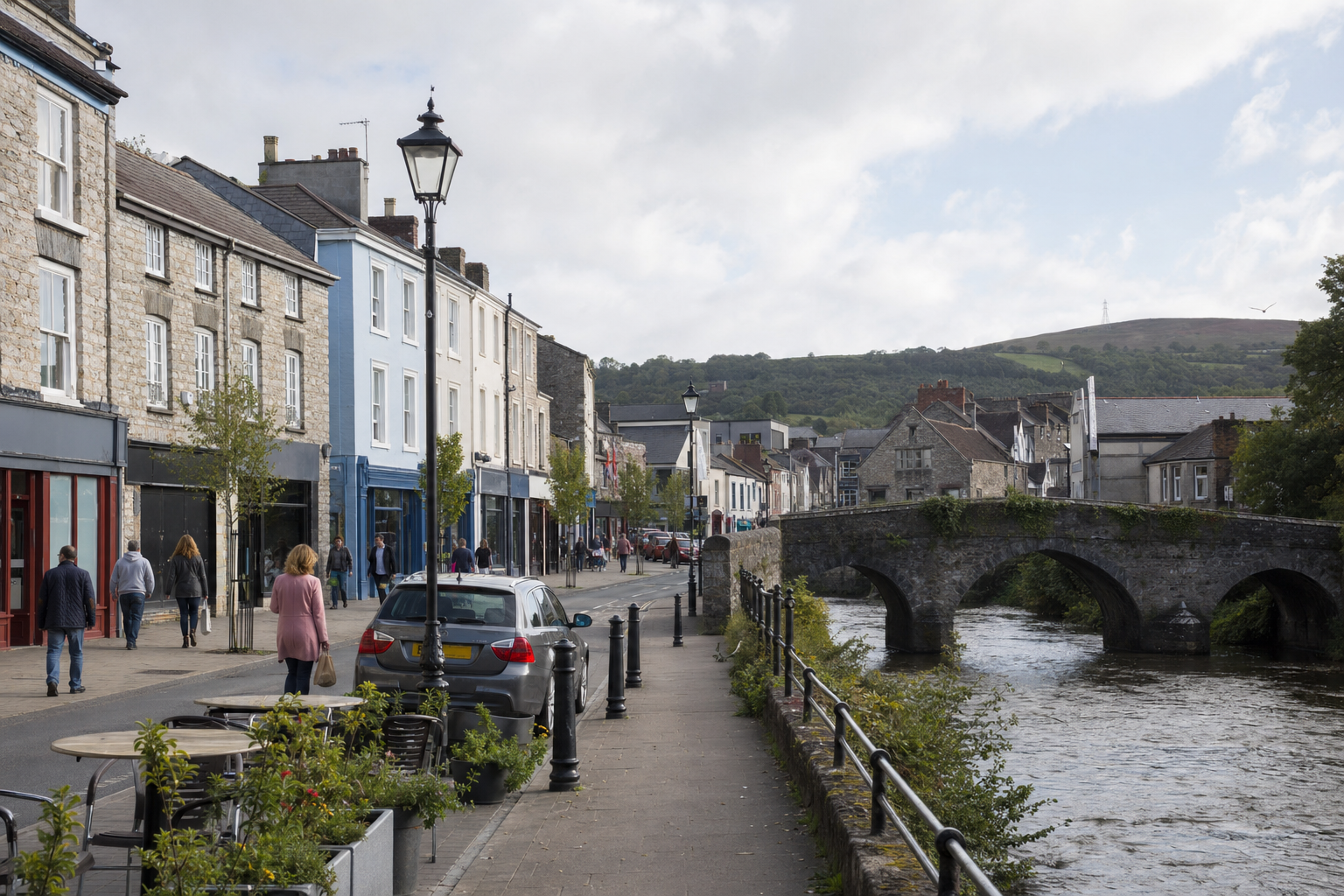 A riverside street with people walking beside historic buildings and an old stone bridge in the background.