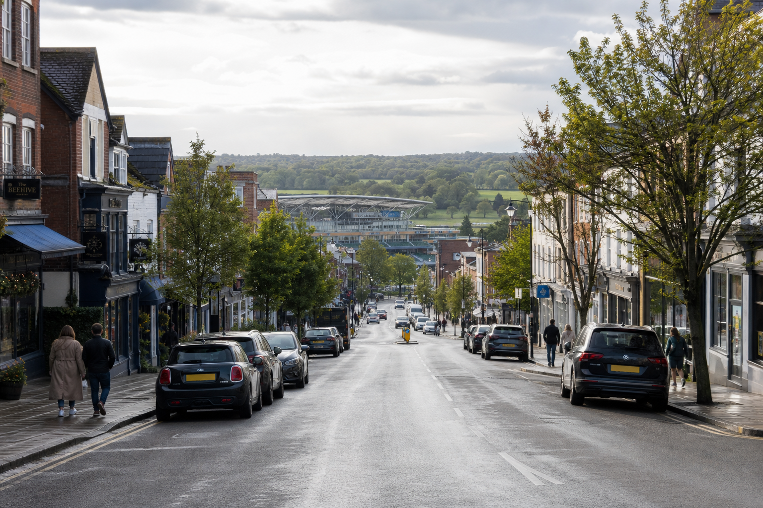 A scenic street lined with parked cars and shops, leading toward a stadium and lush green hills.