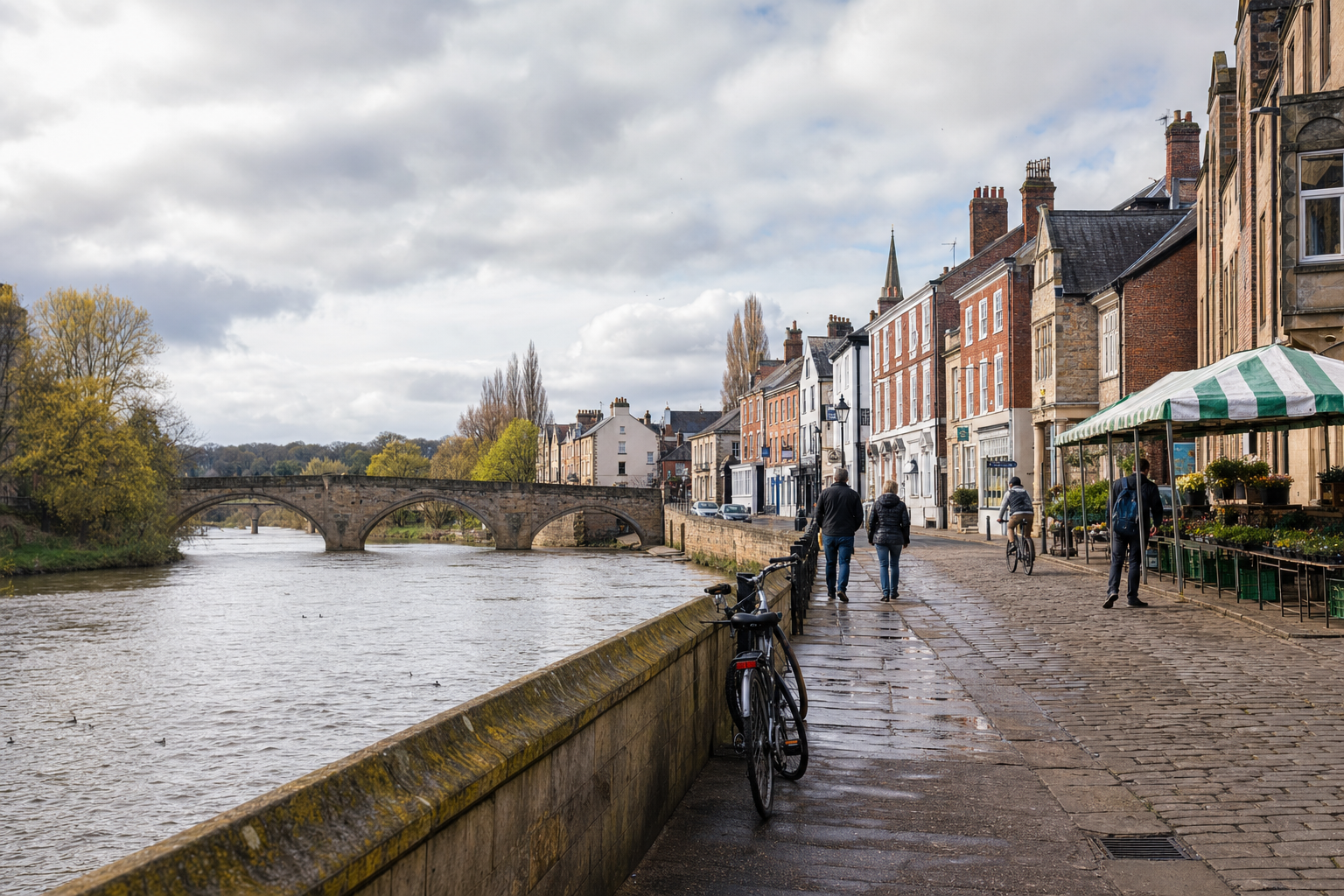 Riverside scene with people walking by colourful buildings and a stone bridge under a cloudy sky.