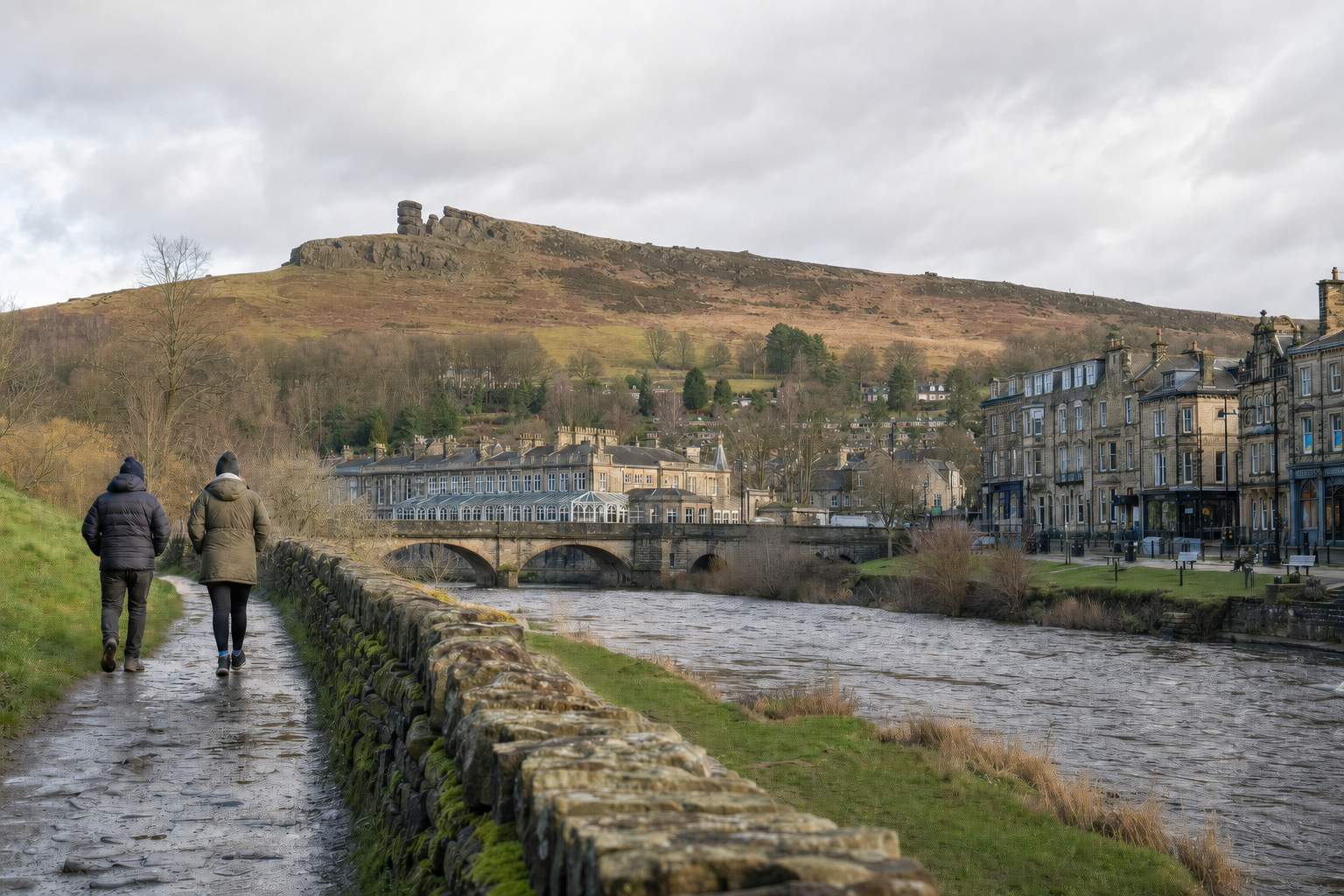 Two people walk along a riverside path in a quaint town with a hill and rocky outcrop in the background.