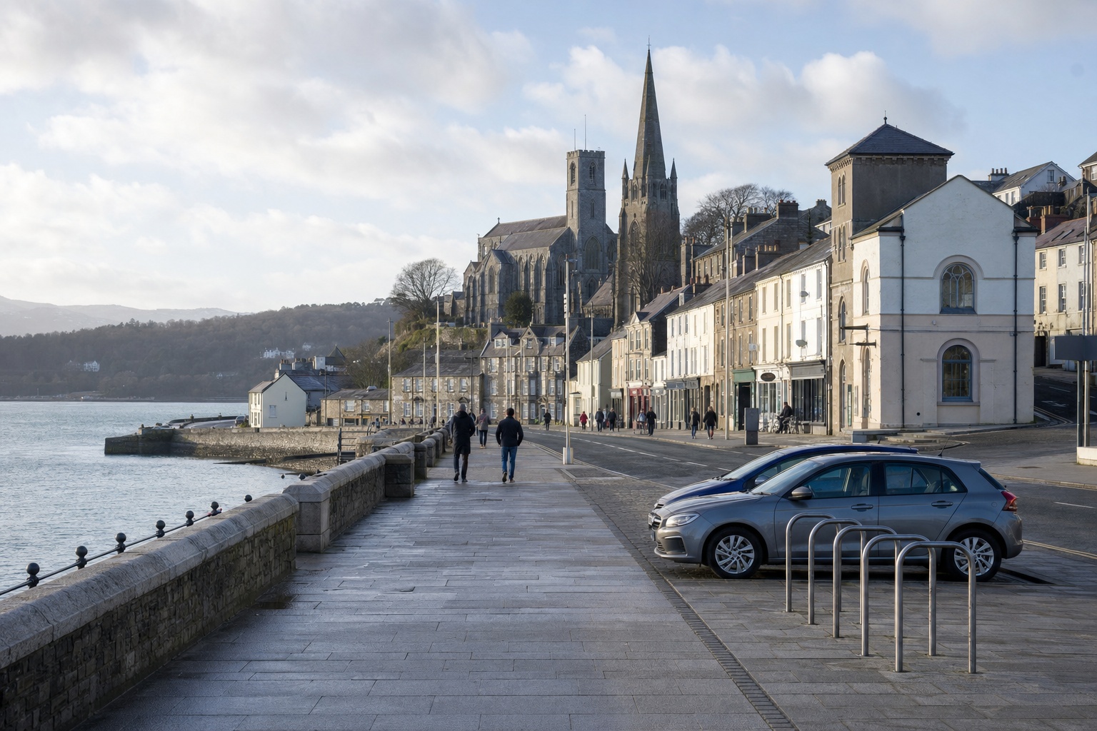 Coastal town street with people walking, cars parked, and a large stone church in the background under a cloudy sky.