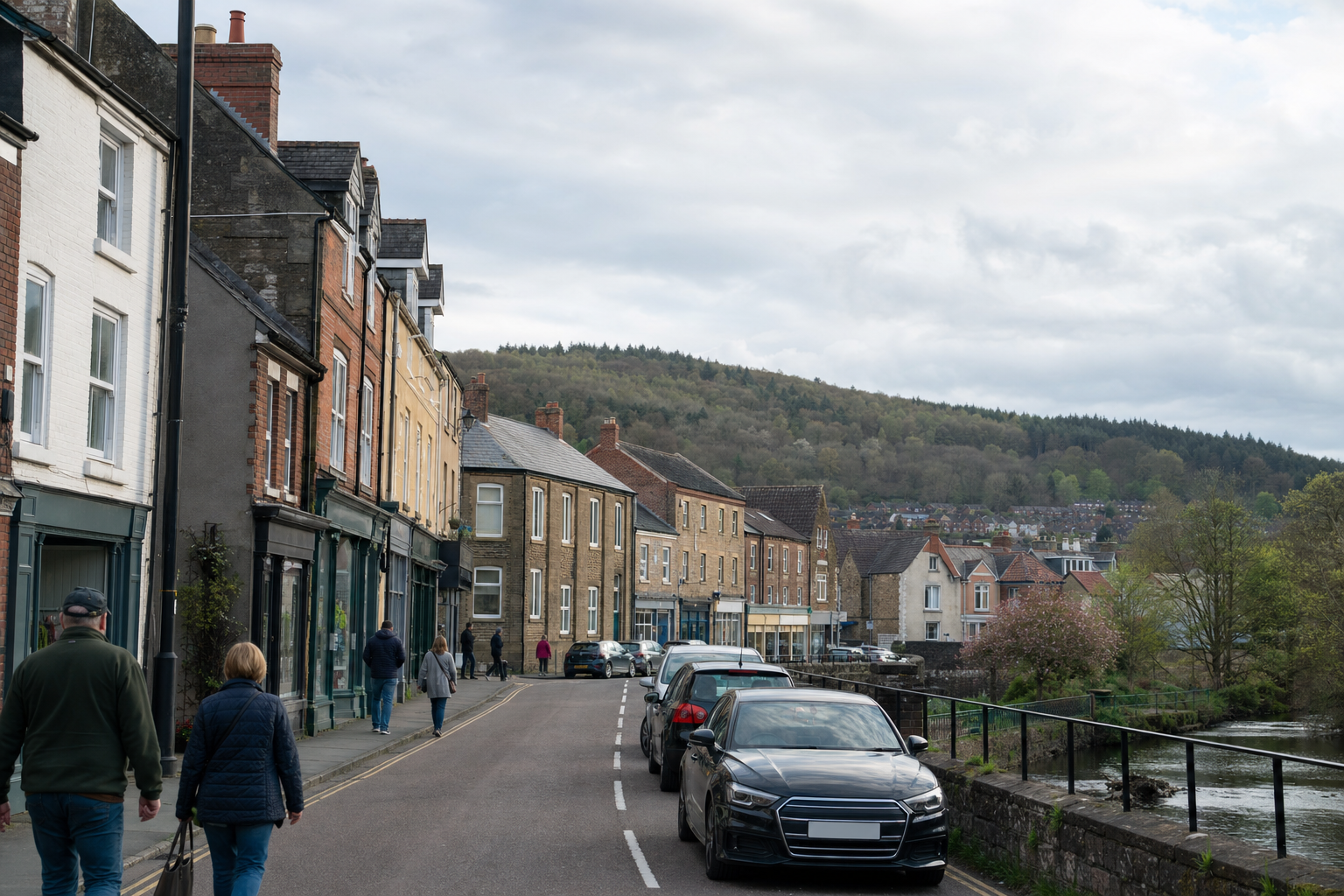 A quiet street lined with terraced houses in a small village, with parked cars and people walking by a river, set against a wooded hillside.
