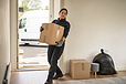 A woman in work gloves carries a cardboard box into a house, with a delivery van parked outside.