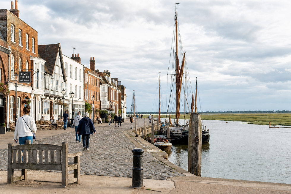 People strolling along a cobbled riverfront street with historic buildings and moored sailboats in the background.