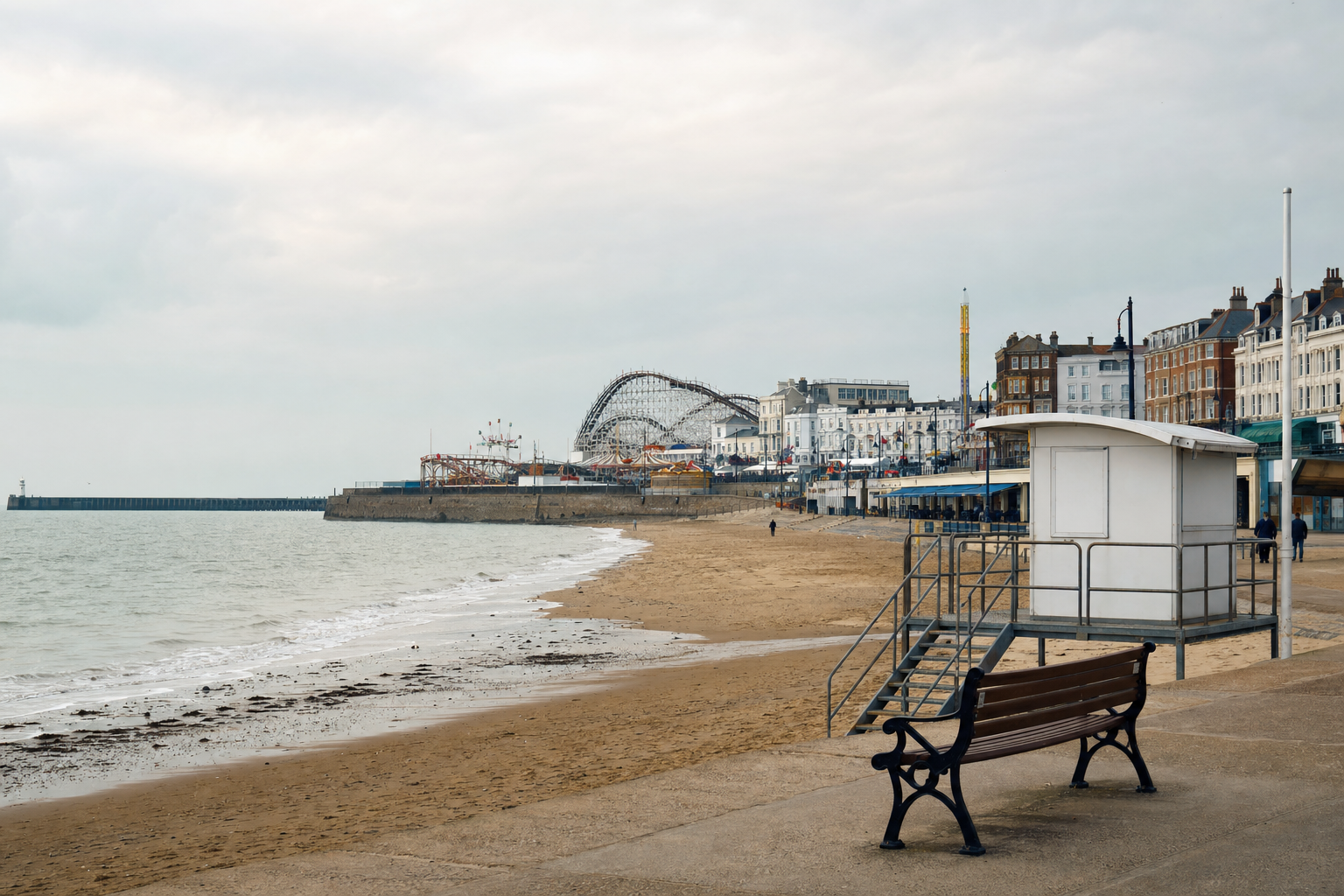 A seaside promenade with a bench overlooks a sandy beach and amusement park against a cloudy sky.