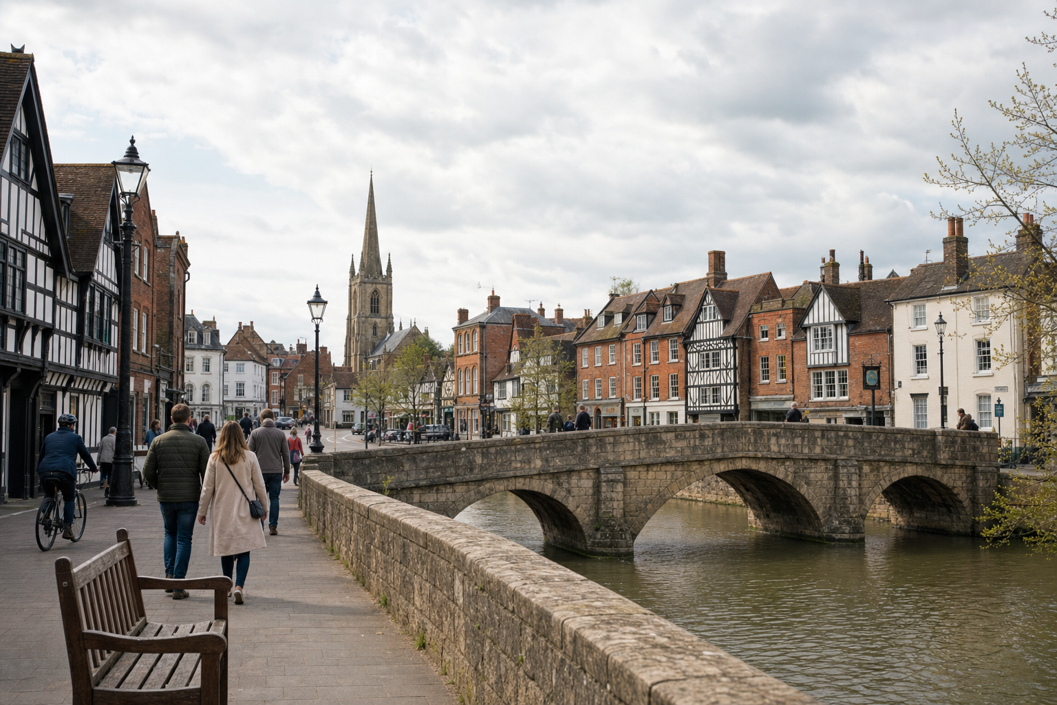 People walking by a river with historic buildings and a church spire in the background.