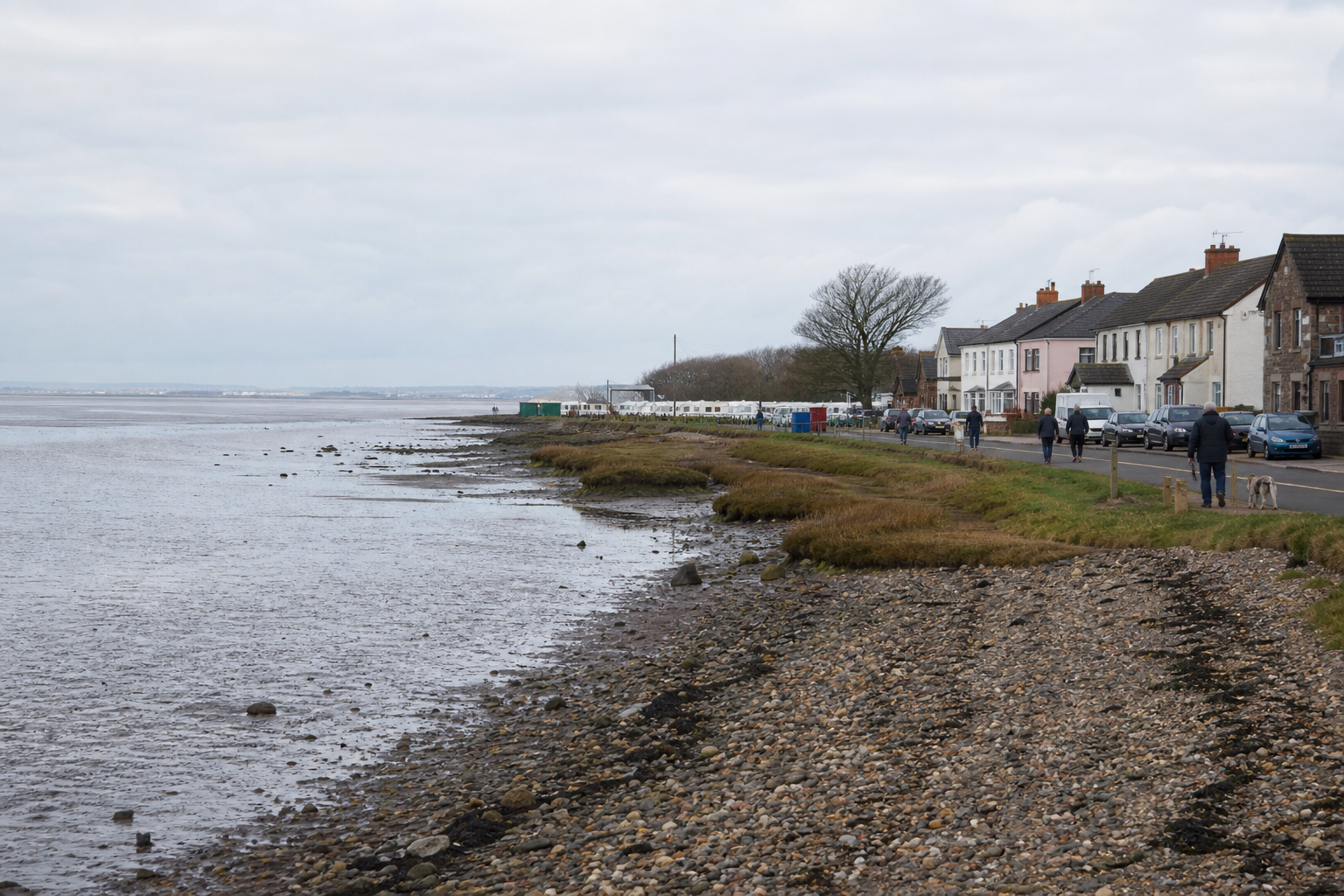 Coastal village with pebble beach, grass, and people walking along the shoreline road.