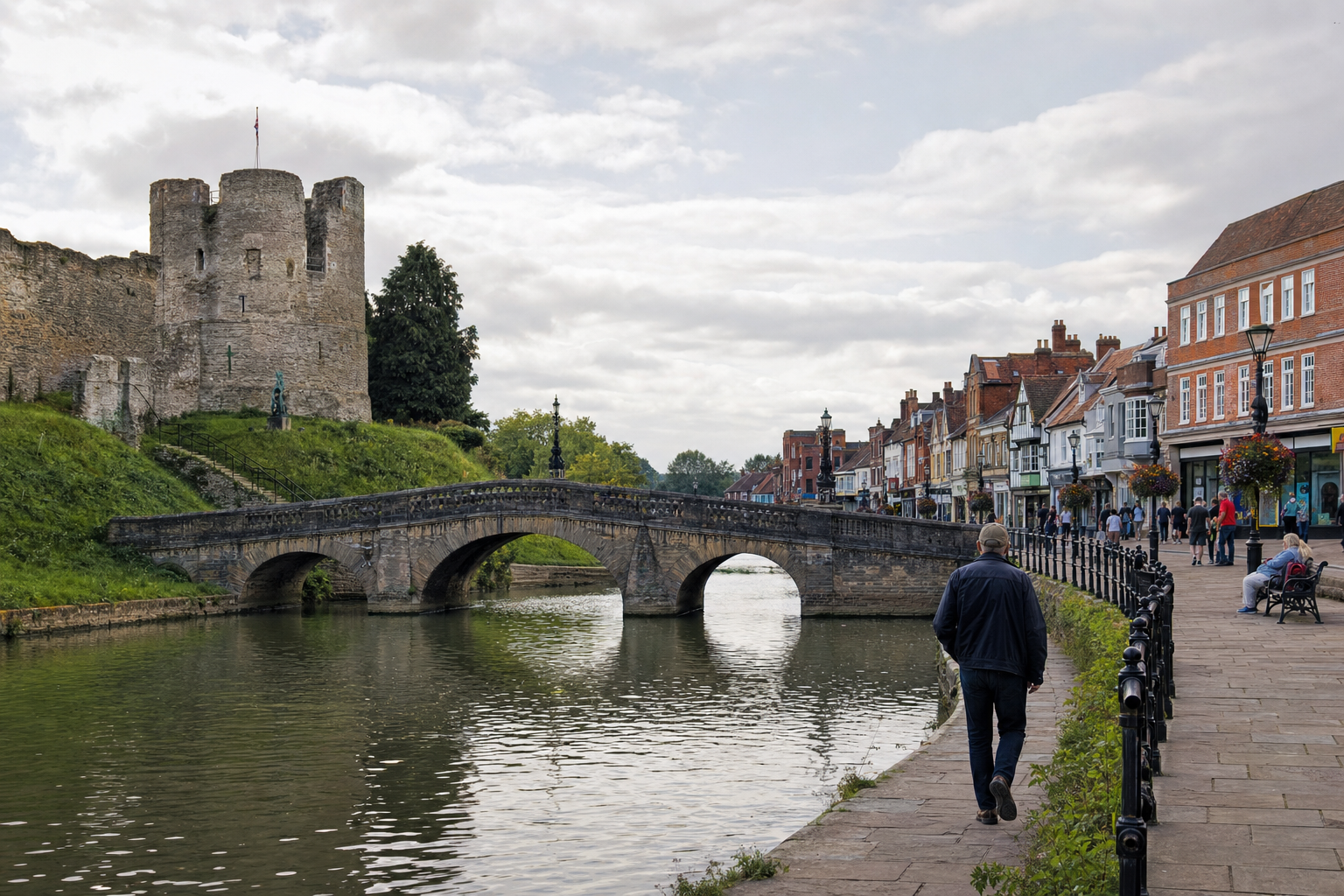 A historic stone bridge spans a river beside a castle and a row of colourful buildings, with people walking along the pathway.