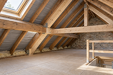 Spacious wooden loft with exposed beams and insulation, featuring a skylight and unfinished flooring.