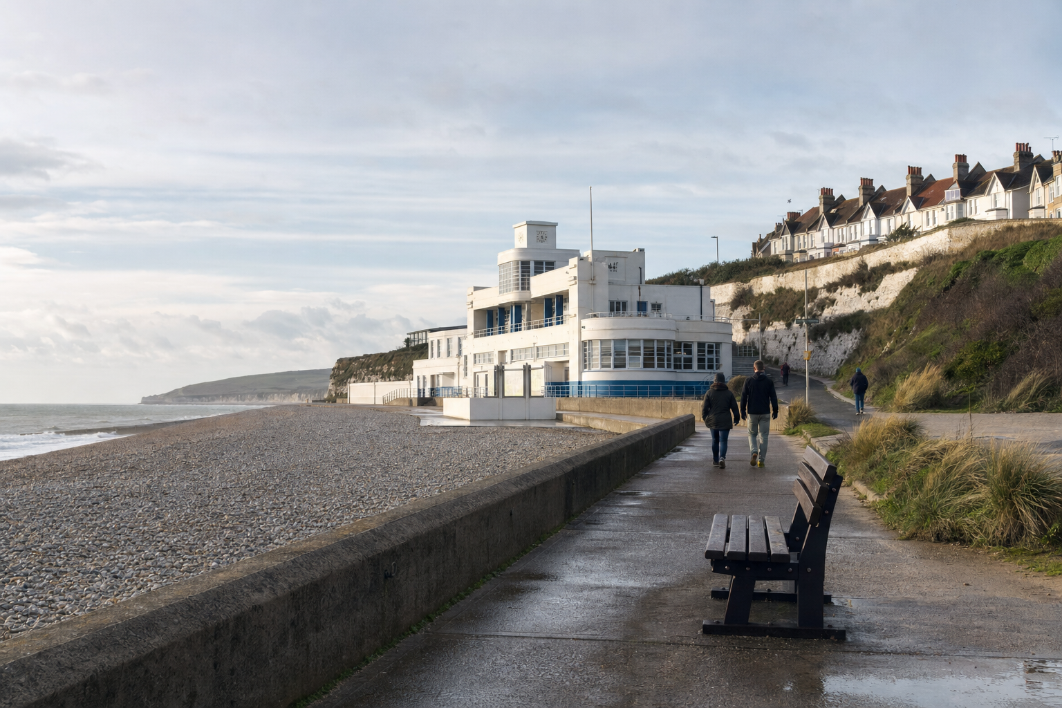 Two people walking along a seaside promenade near an art deco building on a cloudy day.
