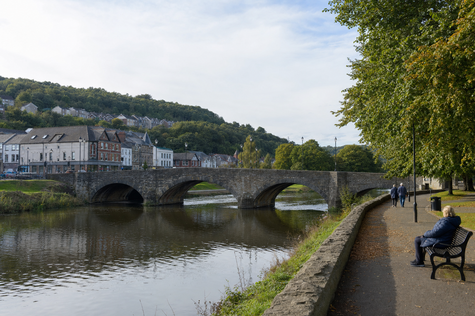Stone bridge over a river with people walking along a tree-lined path on a clear day.