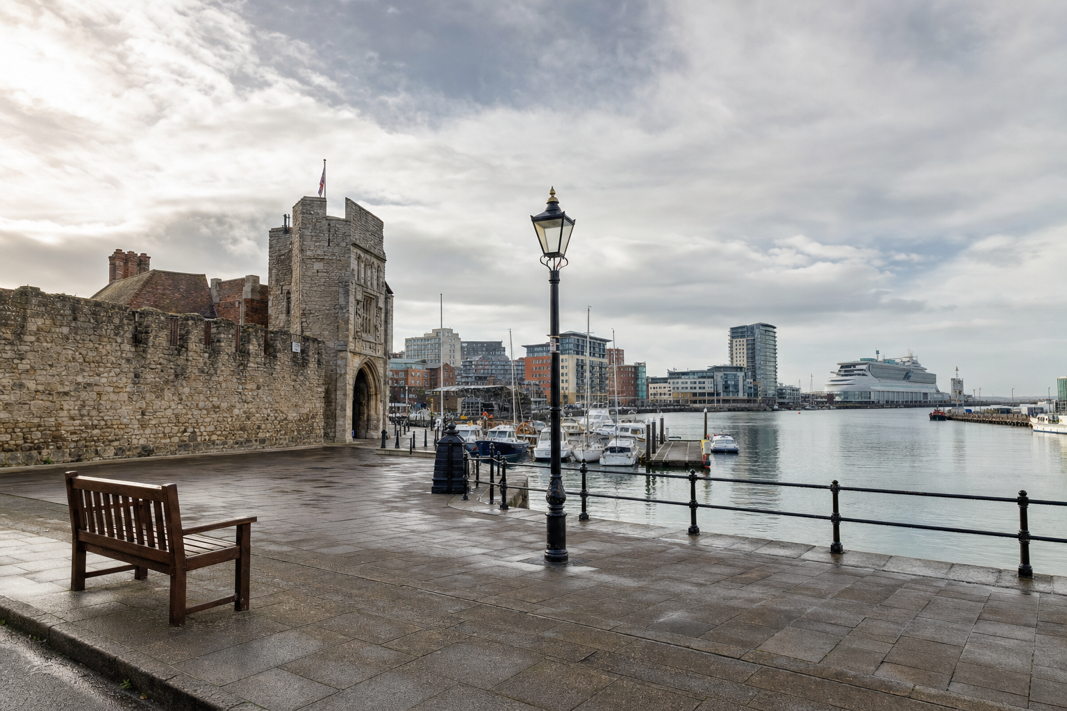 Historic stone gatehouse and adjacent harbour with modern buildings and moored boats under a cloudy sky.