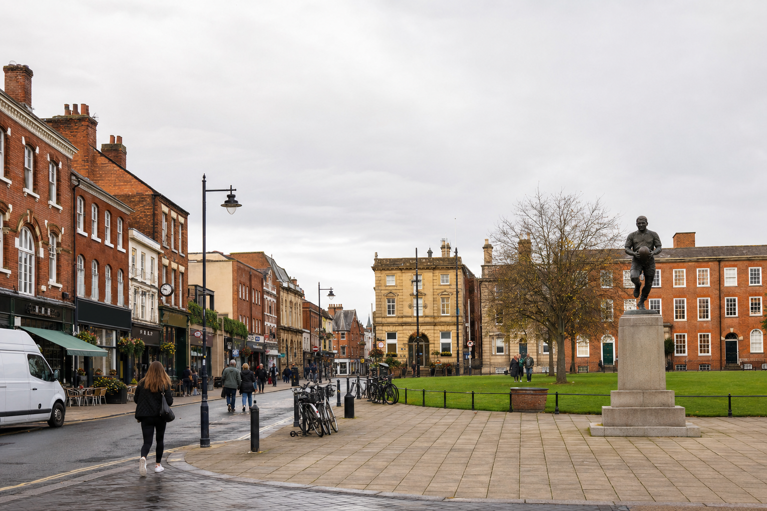 A statue stands prominently in a historic town square surrounded by red brick buildings and people walking.