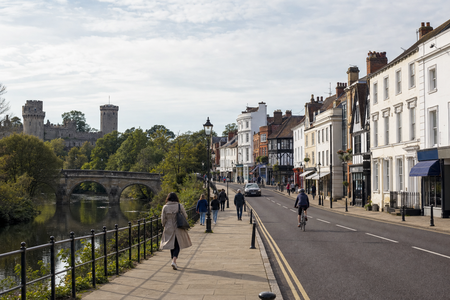 People walking along a riverside pavement with historic buildings and a castle in the background.