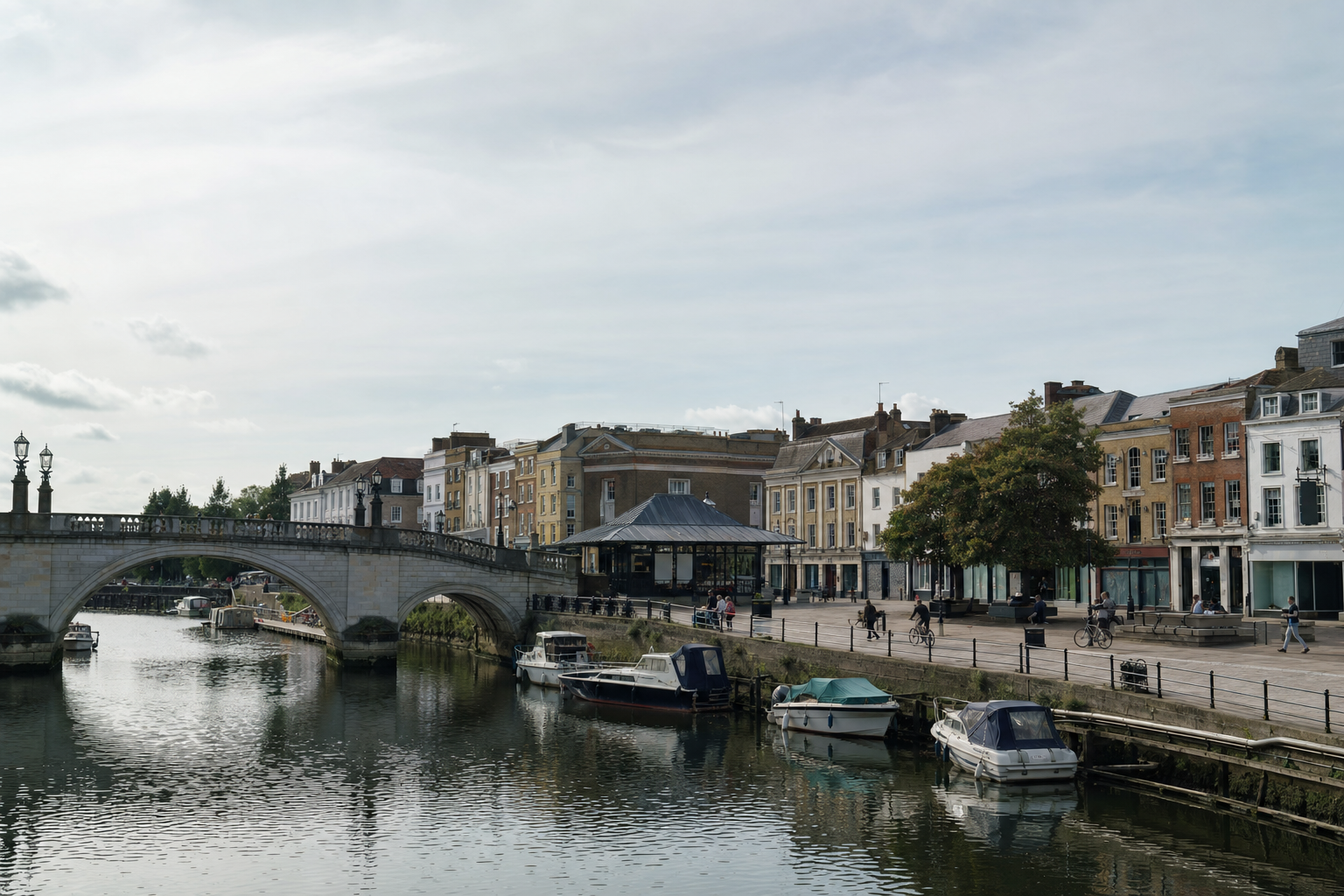 Stone bridge over a river with moored boats and colourful buildings lining the promenade under a partly cloudy sky.