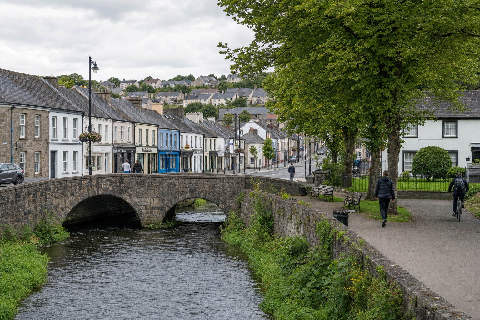 A stone bridge spans a river flowing through a charming village with historic buildings and trees along the path.