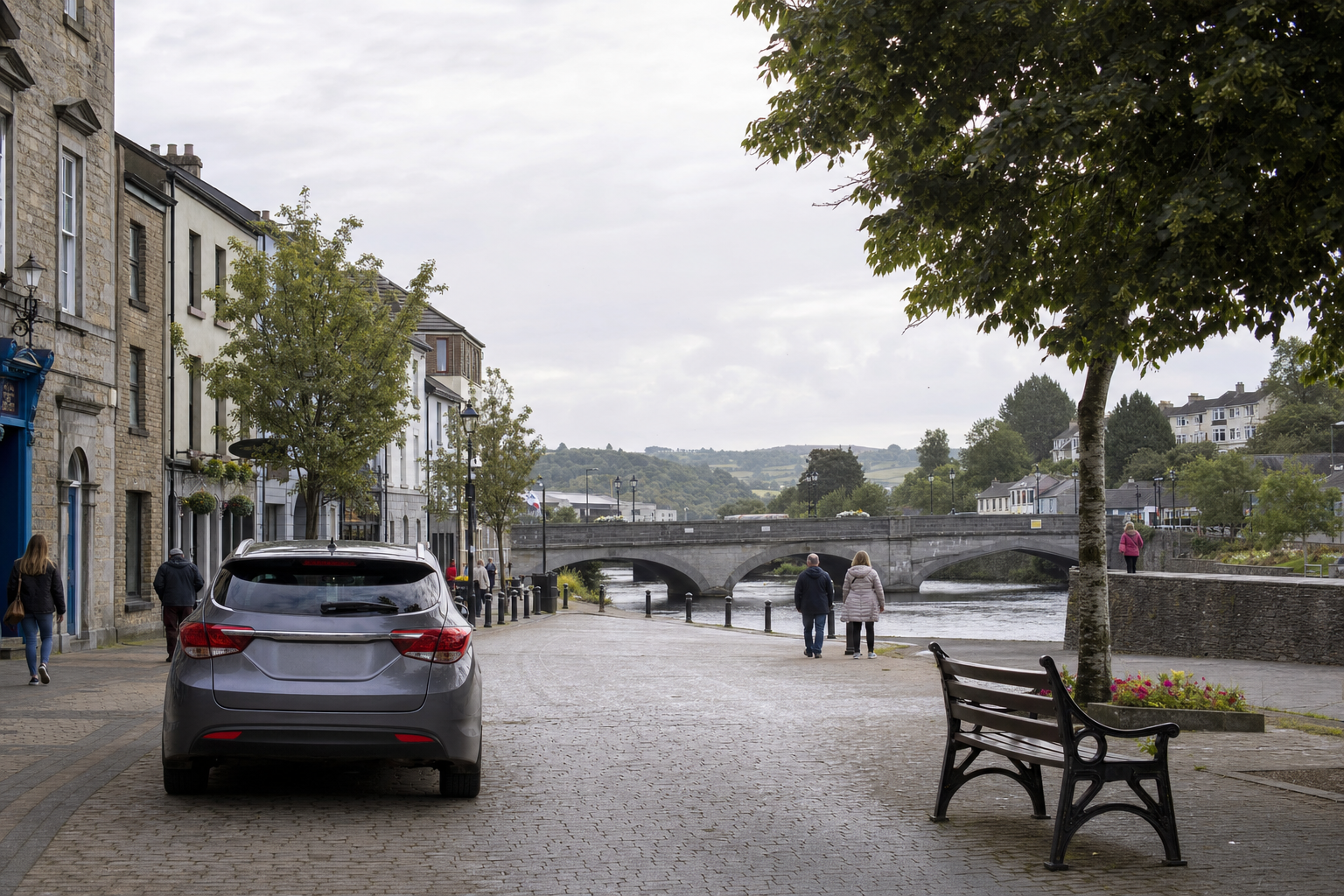 A quaint riverside street with a car, pedestrians, and a stone bridge in the distance under a cloudy sky.