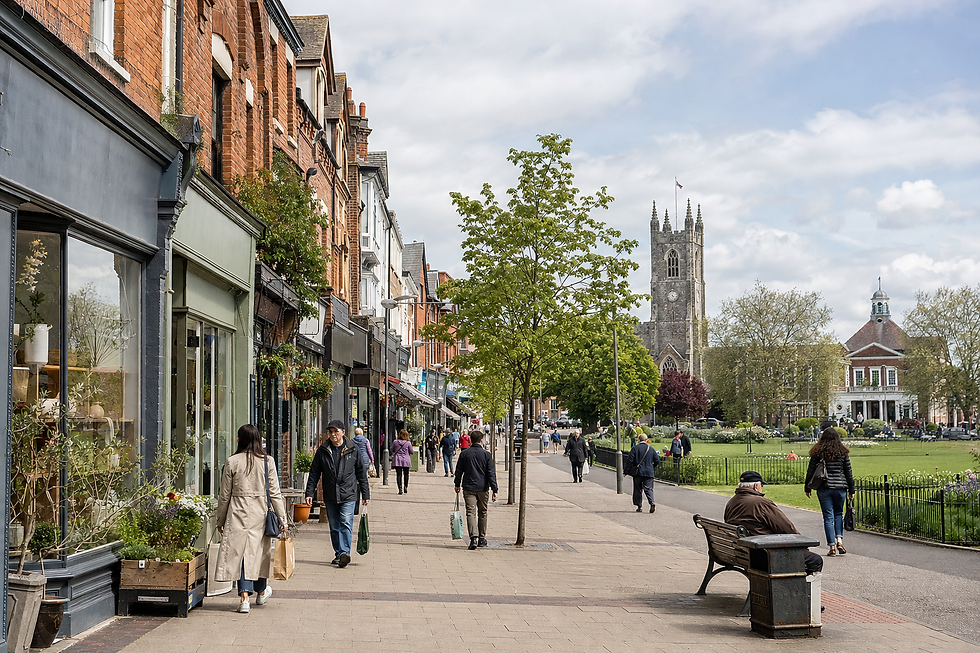 A bustling high street with people walking and shops lining the pavement, leading to a historic church and park in the background.