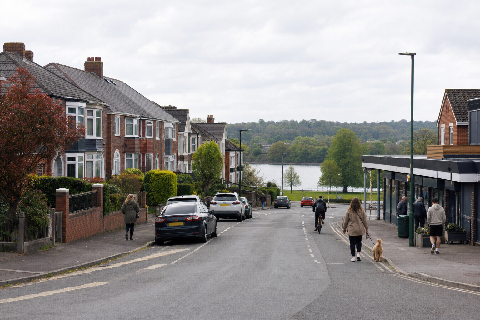 A suburban street with terraced houses, cars parked along the roadside, and people walking and cycling towards a lake.