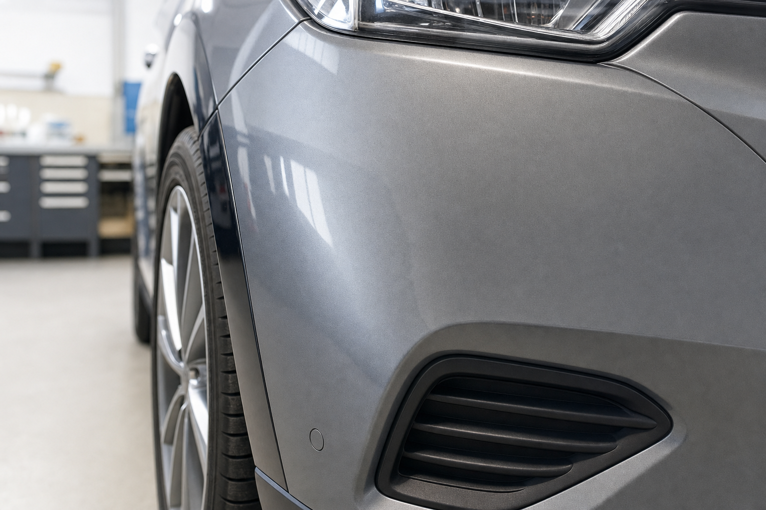 Close-up of a grey car's front side with alloy wheel and vent in a garage setting.