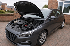 A grey hatchback parked on a driveway with the bonnet open, revealing the engine compartment.