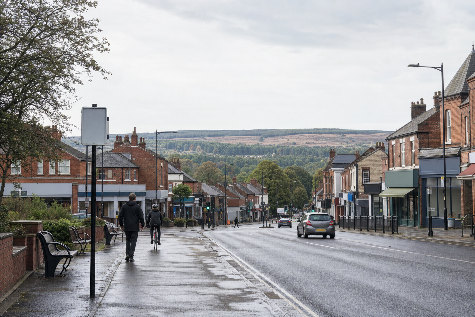 A quiet street in a small town with a cyclist and pedestrian on a rainy day, lined with brick buildings and trees in the distance.