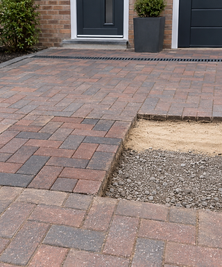 Brick driveway with a missing section and stacked bricks beside, in front of a modern house entrance.