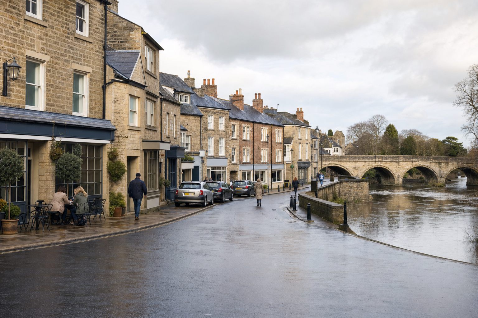 Quaint British street with stone buildings, a few pedestrians, and a historic stone bridge over a river.
