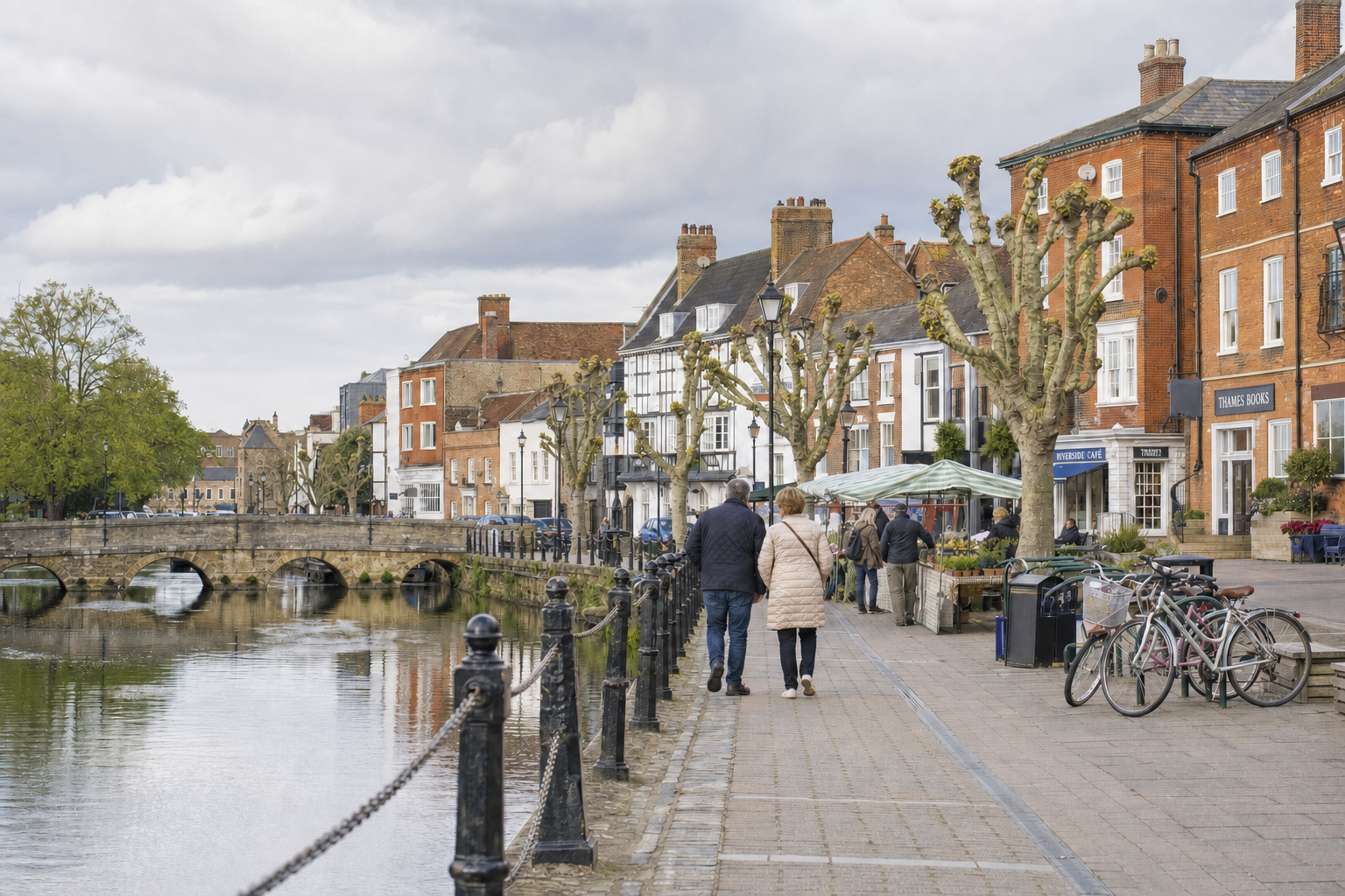 A couple walks by a river with a stone bridge and picturesque shops in the background.